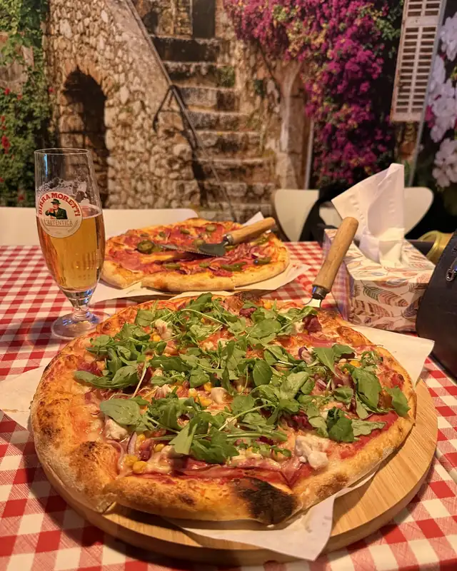 Pizza z rukolą na drewnianej desce, na obrusie w czerwoną kratkę, w tle mural z uliczką